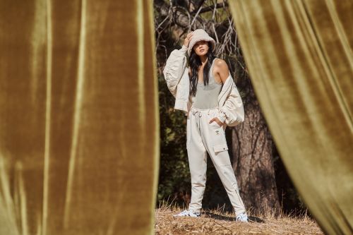 A woman models Adidas apparel in an outdoor setting with trees and golden curtain-like material flanking the sides of the frame.