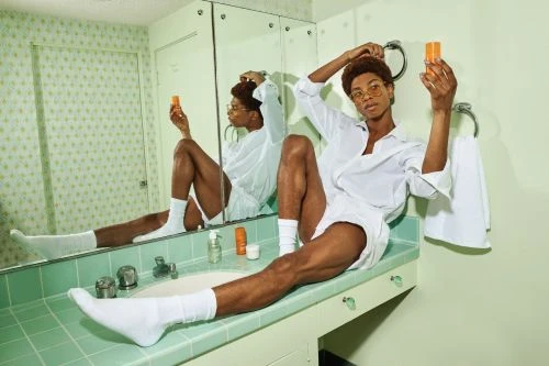 Person sits on bathroom counter in white shirt, shorts, socks and glasses holding orange cosmetic bottle near mirror in a bathroom setting.