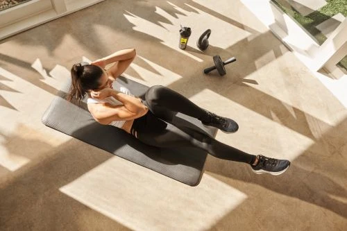 Athletic woman in black leggings and white top performs crunches on a mat indoors with workout gear nearby and bright natural lighting.