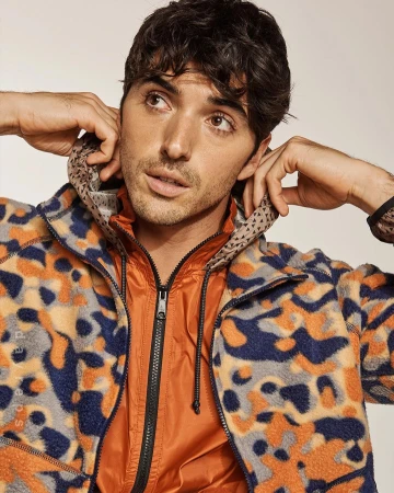 Adult male wearing a patterned jacket with a hood is featured in a portrait shot against a plain background, fashion editorial. 