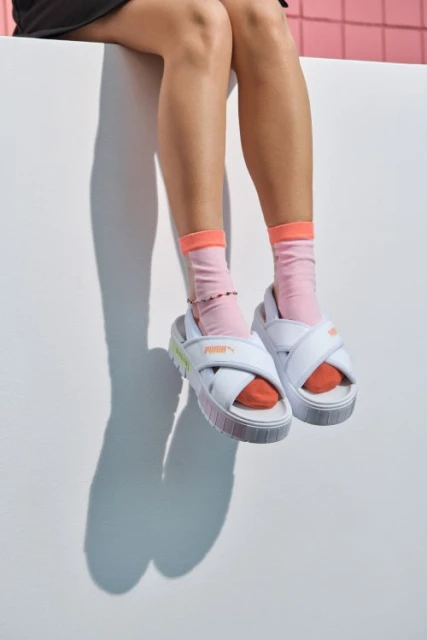 A woman's legs and feet wearing white Puma platform sandals and pink socks are seen sitting on a white wall with a pink background.