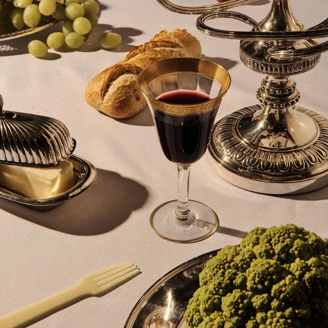 Elegant table setting with wine in a gold-rimmed glass, bread, grapes, butter dish, plastic fork, and romanesco broccoli on a white tablecloth.