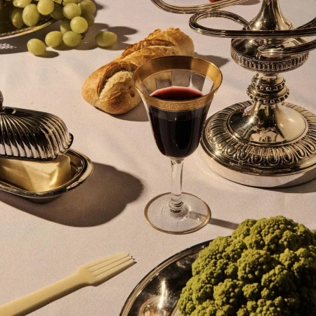 Elegant table setting with wine in a gold-rimmed glass, bread, grapes, butter dish, plastic fork, and romanesco broccoli on a white tablecloth.