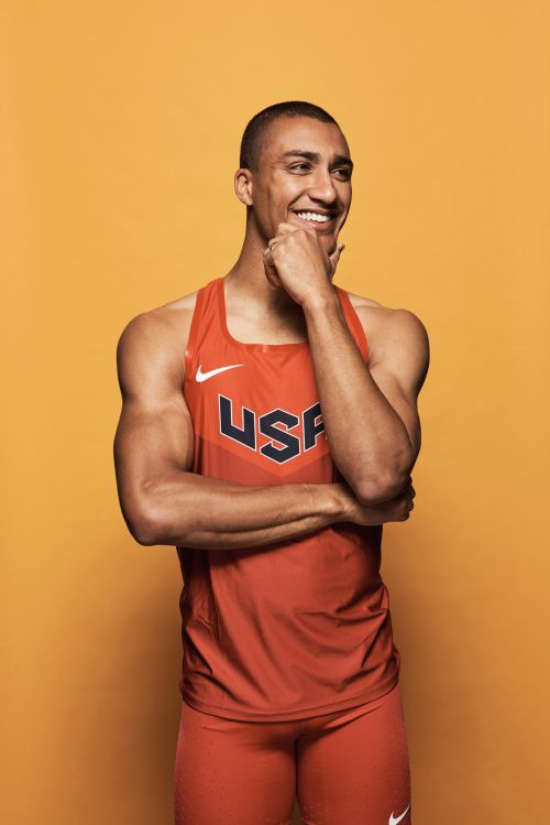 Smiling male athlete in a red USA-branded athletic top, with hand to chin. The image is set against a solid yellow backdrop.