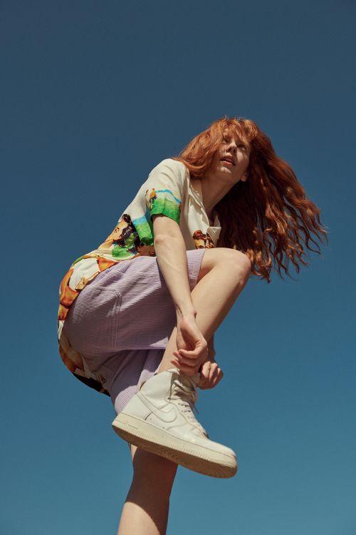 A redhead woman in a patterned button-down shirt and lilac shorts tying her sneaker against a clear blue sky backdrop.
