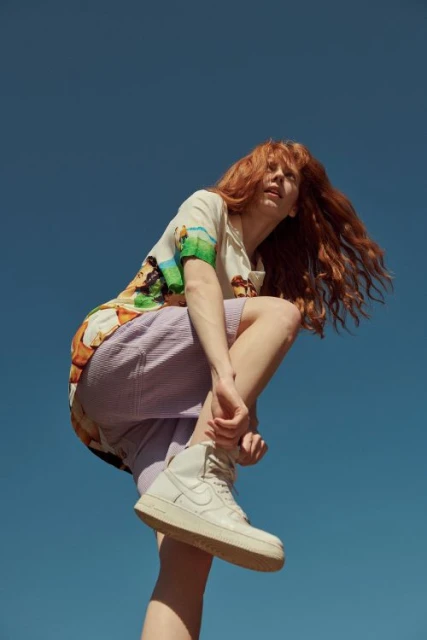 A redhead woman in a patterned button-down shirt and lilac shorts tying her sneaker against a clear blue sky backdrop.