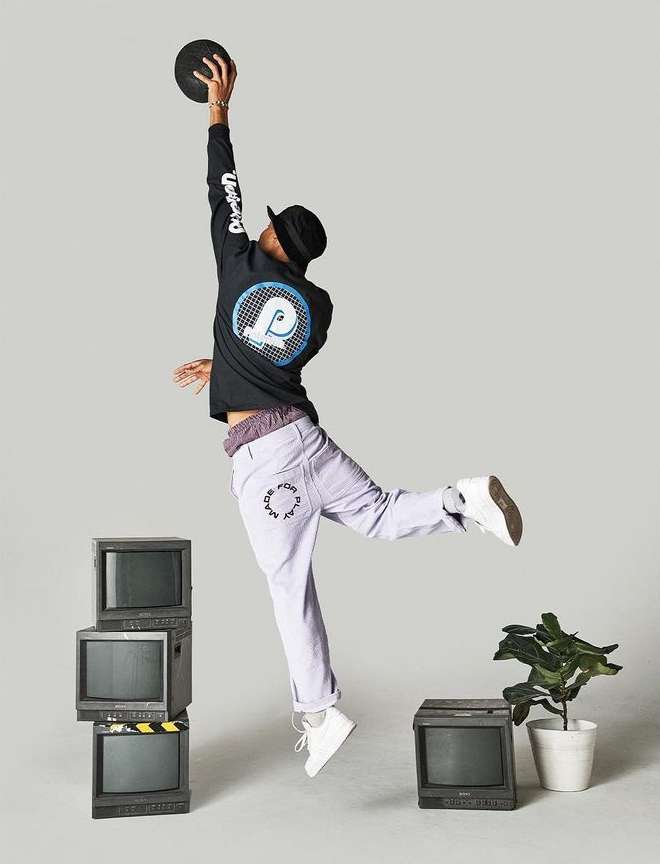 A man jumps in the air toward a black disc, wearing a black hat, black long-sleeve shirt, light purple pants, and white sneakers, in a photo studio.