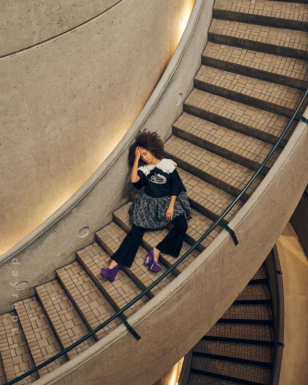 Woman in a black top, gray skirt, and purple platform shoes sits on a concrete staircase with a curved railing in an indoor space.