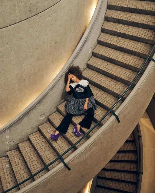 Woman in a black top, gray skirt, and purple platform shoes sits on a concrete staircase with a curved railing in an indoor space.