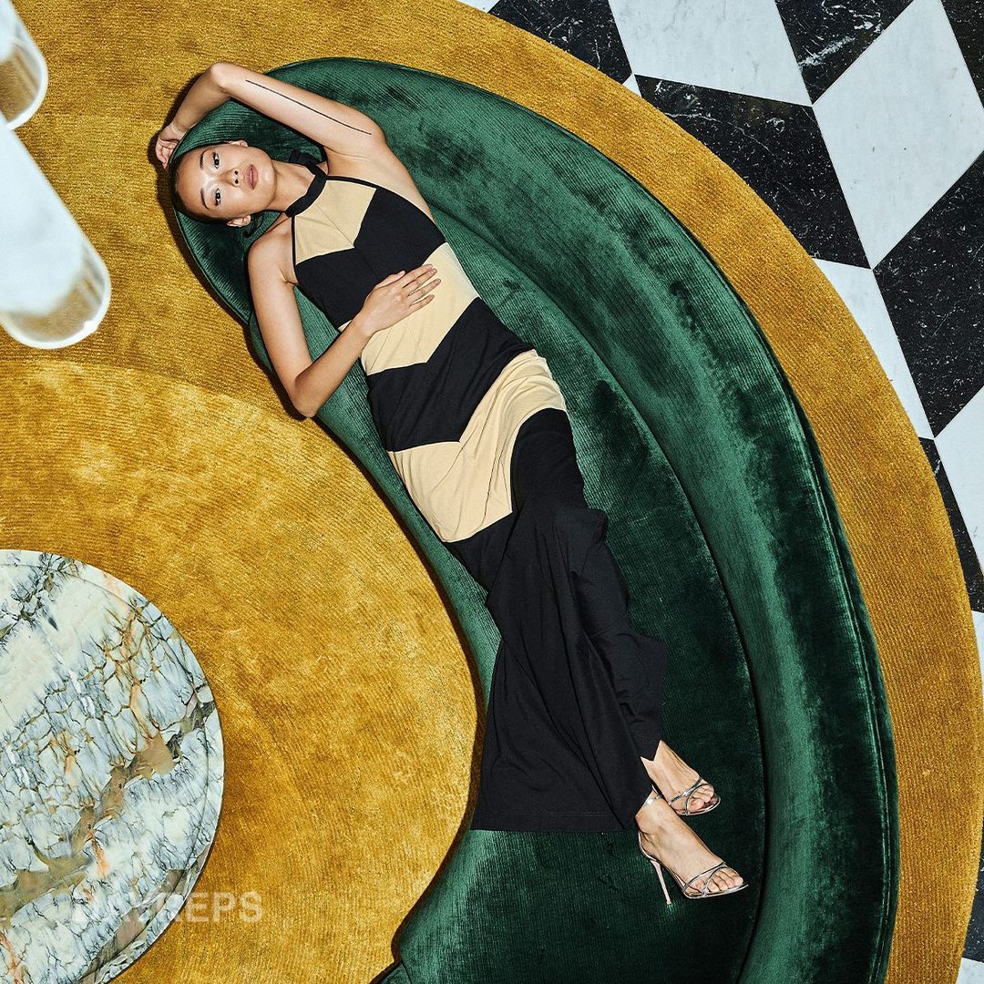 Overhead shot of a young woman in a chevron-pattern halter dress reclining on a green velvet couch, with marble and gold accents.