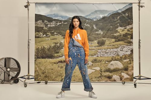 Full-length studio shot of a young Asian woman wearing an orange hoodie and paint-splattered denim overalls, posing in front of a backdrop.