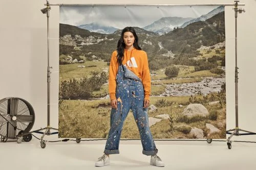 Full-length studio shot of a young Asian woman wearing an orange hoodie and paint-splattered denim overalls, posing in front of a backdrop.