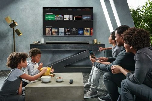 A family of five watches television together in a modern living room setting, sharing snacks and using a smartphone while seated on a gray sofa.
