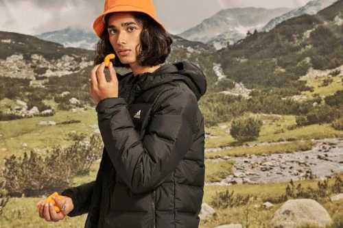 Man in a black puffer jacket and orange hat standing in a mountain landscape, eating an orange snack, with a backdrop of mountains.