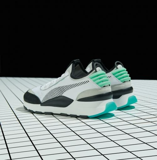 A pair of white Puma sneakers with black and teal accents are angled on a white grid floor against a black background, product shot.