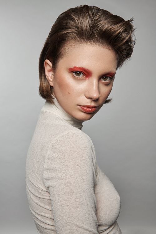 Close-up portrait of a young woman with short brown hair, neutral turtleneck top, and striking red eye shadow against a grey background.