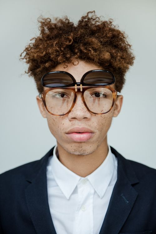 Portrait of young man with curly brown hair wearing tortoiseshell glasses and a dark blazer over a white shirt against a plain background.