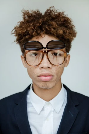 Portrait of young man with curly brown hair wearing tortoiseshell glasses and a dark blazer over a white shirt against a plain background.