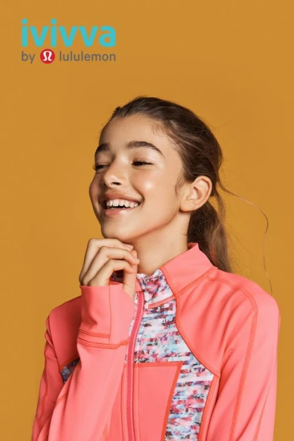 A smiling tween girl in a coral jacket poses against a warm yellow backdrop, showcasing athletic fashion by Ivivva by Lululemon.
