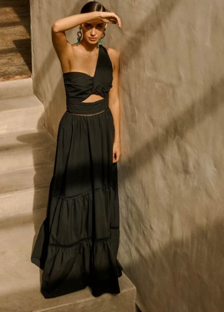 Fashionable woman in a one-shoulder black dress poses on concrete stairs next to textured wall in a natural light setting.
