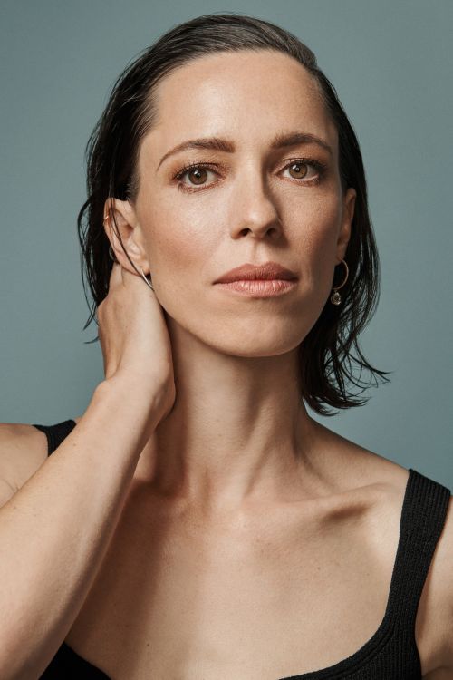 Close-up portrait of an adult woman with slicked-back dark hair wearing a black tank top against a solid blue background.