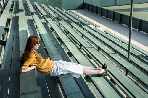 Redhead woman in a yellow sweater and white culottes lounges on green wooden bleachers near a tennis court on a sunny day.