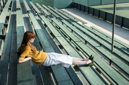 Redhead woman in a yellow sweater and white culottes lounges on green wooden bleachers near a tennis court on a sunny day.
