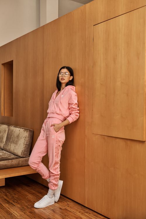A slender woman wearing a pink Adidas sweatsuit leans against a wooden wall in a lifestyle shot inside, showcasing fashion and style.