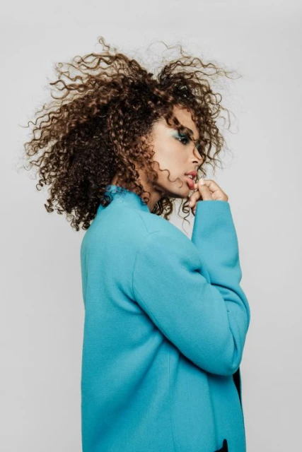 Portrait of a curly-haired woman wearing a turquoise jacket. Close-up shot of woman in stylish attire posing against a light backdrop.