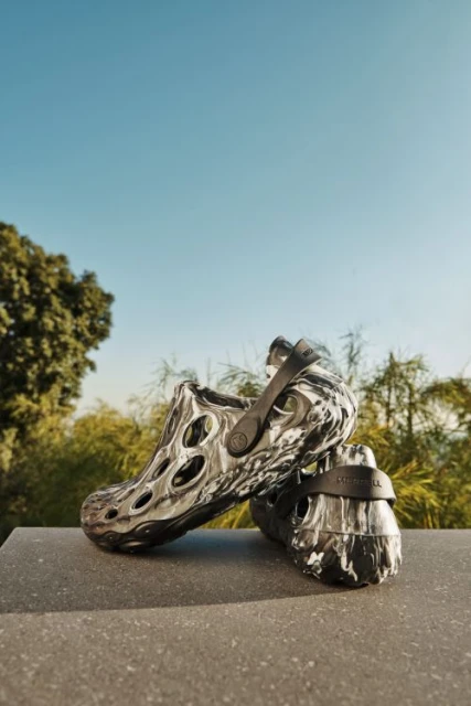 Two black and white Merrell clogs on a stone surface against a natural outdoor backdrop of trees and a clear blue sky.