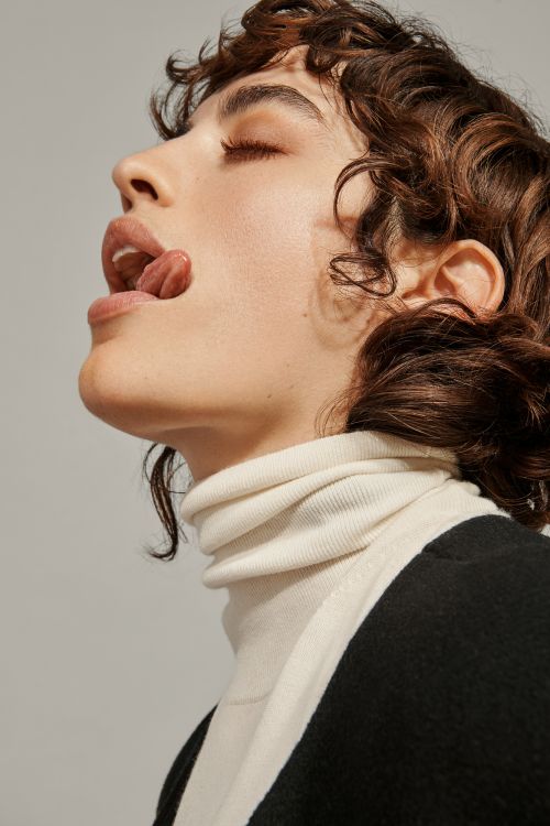 Portrait of a woman with curly brown hair and closed eyes sticking her tongue out while wearing a white turtleneck and black jacket.
