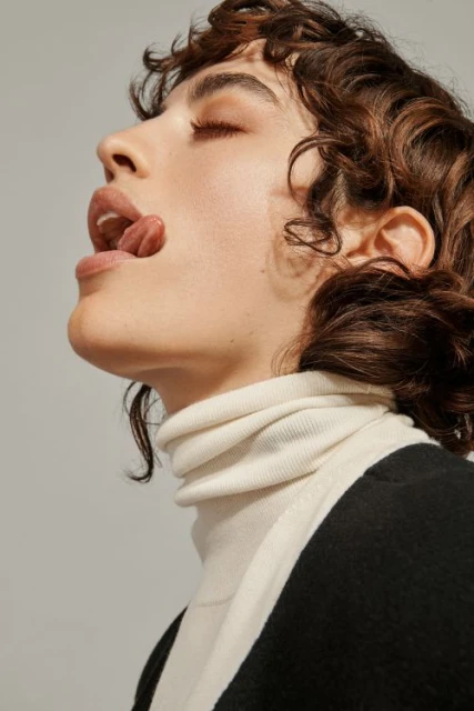 Portrait of a woman with curly brown hair and closed eyes sticking her tongue out while wearing a white turtleneck and black jacket.