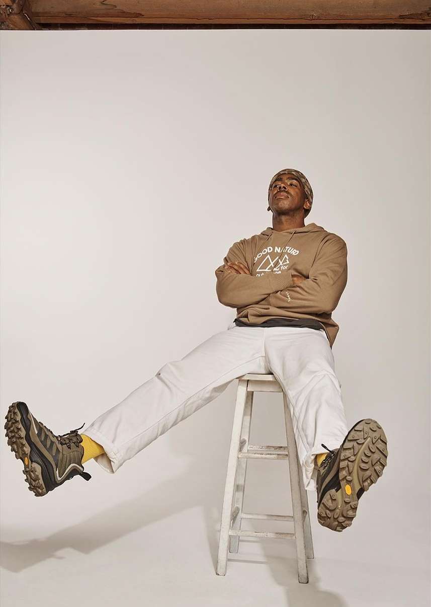 Man in hoodie and white pants sits on a stool in a photo studio, legs extended, wearing hiking boots in a studio shot.