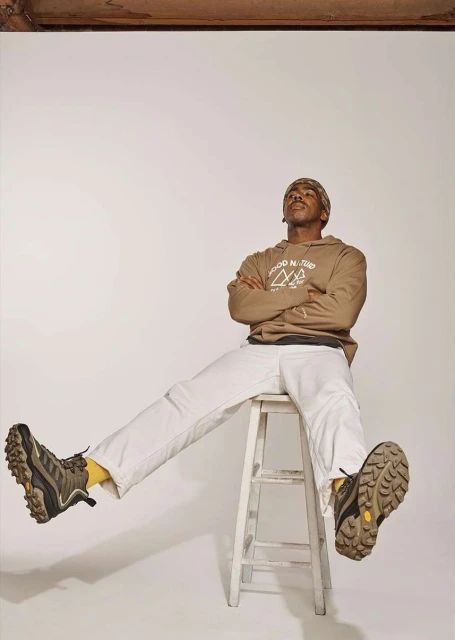 Man in hoodie and white pants sits on a stool in a photo studio, legs extended, wearing hiking boots in a studio shot.