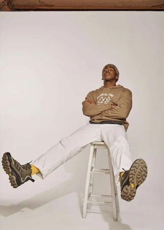 Man in hoodie and white pants sits on a stool in a photo studio, legs extended, wearing hiking boots in a studio shot.