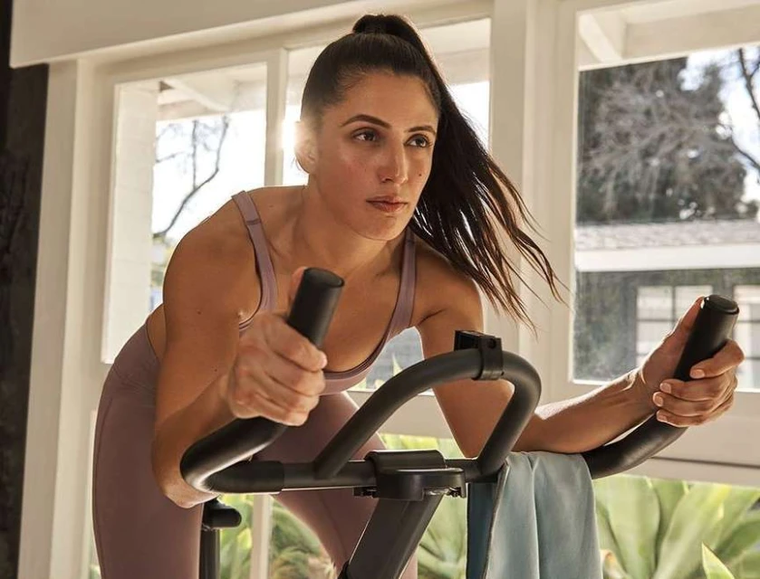 Woman works out on an exercise bike in a brightly lit room with a window and plant in background, wearing athletic clothing.