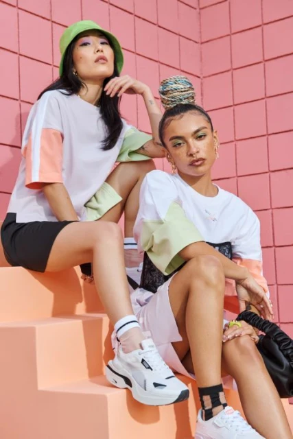 Two women pose on stairs in color-blocked shirts and athletic wear, featuring a pink tiled wall, white sneakers, and coordinated styling.