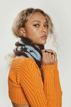 Close-up portrait of a woman wearing an orange ribbed sweater and a dark brown and blue patterned scarf against a white background.