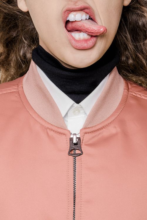 Close-up studio shot of a young fair-skinned woman sticking her tongue out, wearing a pink bomber jacket, black turtleneck, and white collared shirt.