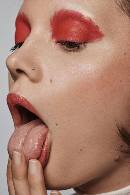 Close-up studio shot of a young adult female with red eyeshadow and red lipstick, sticking out her tongue, and touching her lips.