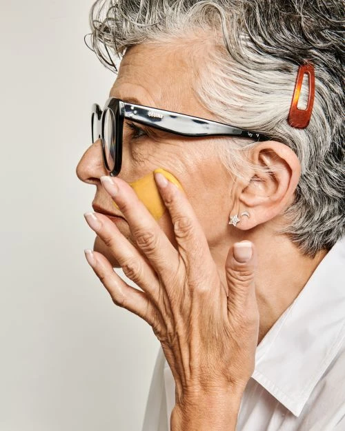 Elderly woman applying makeup to her face in a close-up studio shot. The image features grey hair and eyeglasses.