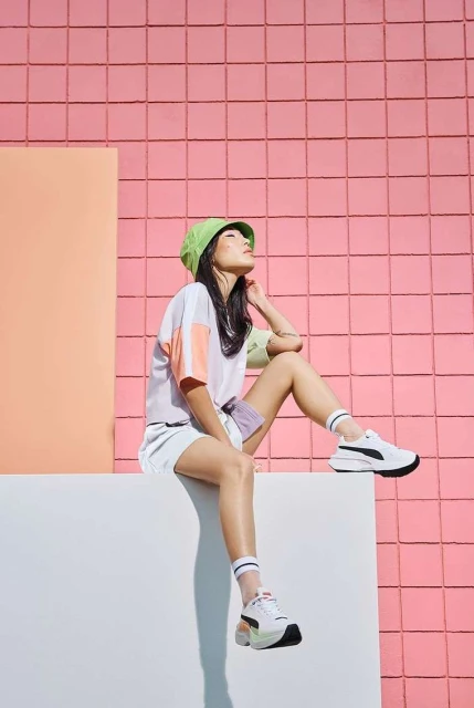 Young woman in stylish athleisure wear posing outdoors against a pink tiled wall, wearing sneakers, a green bucket hat, and coordinated separates.