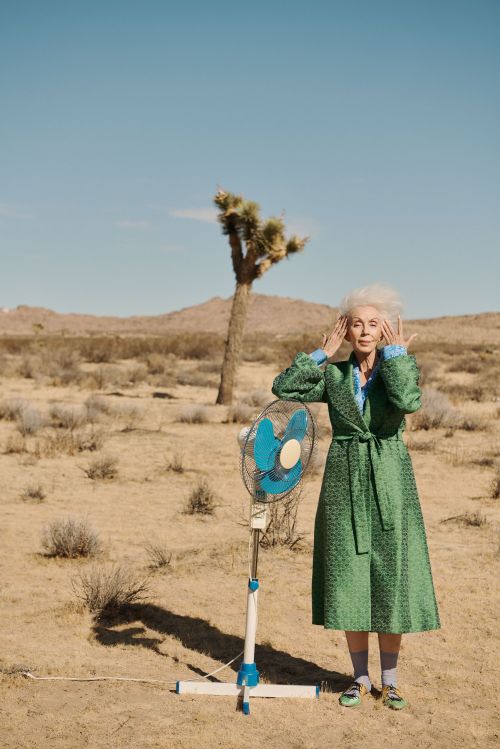 Elderly woman in robe poses next to fan in desert landscape. She has white hair and hands near her face. Joshua tree in background.
