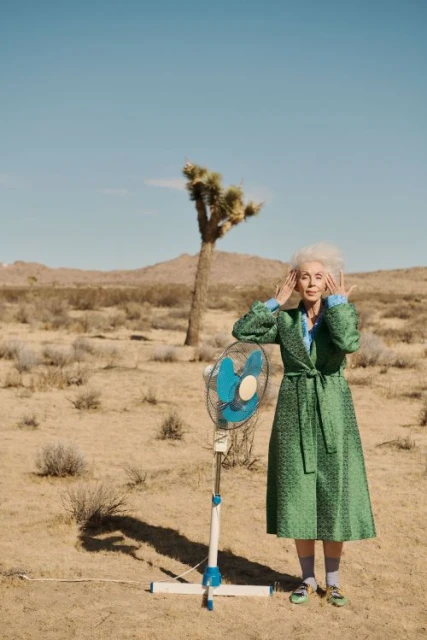 Elderly woman in robe poses next to fan in desert landscape. She has white hair and hands near her face. Joshua tree in background.