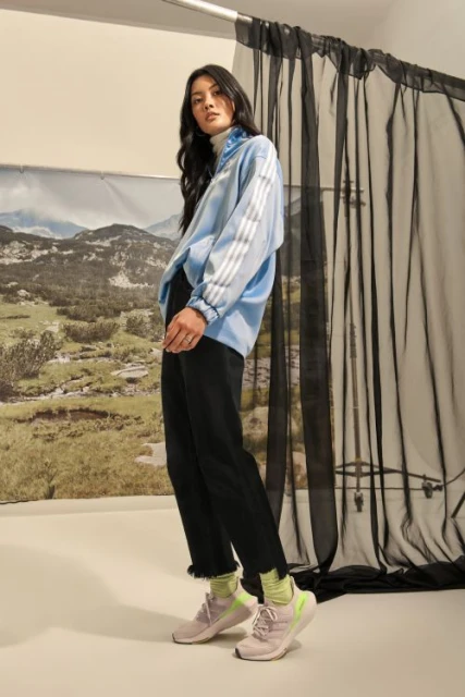 Woman in a light blue athletic jacket and black pants poses in front of a mountain backdrop and black curtains indoors.