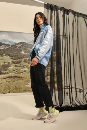 Woman in a light blue athletic jacket and black pants poses in front of a mountain backdrop and black curtains indoors.