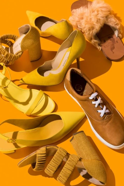 Collection of yellow footwear including high heels, sandals, flats, slippers and sneakers arranged on an orange background in a studio setting.