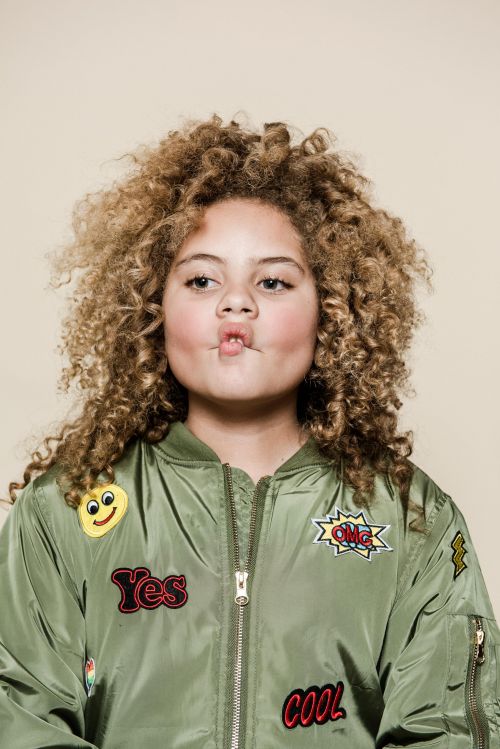 Portrait of a young girl with curly blonde hair wearing a green bomber jacket with patches against a neutral beige background.