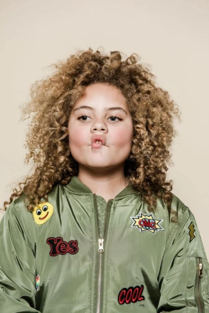 Portrait of a young girl with curly blonde hair wearing a green bomber jacket with patches against a neutral beige background.