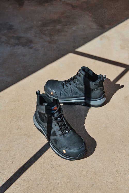Pair of black leather work boots are placed in direct sunlight. The composition is a product shot on concrete flooring.
